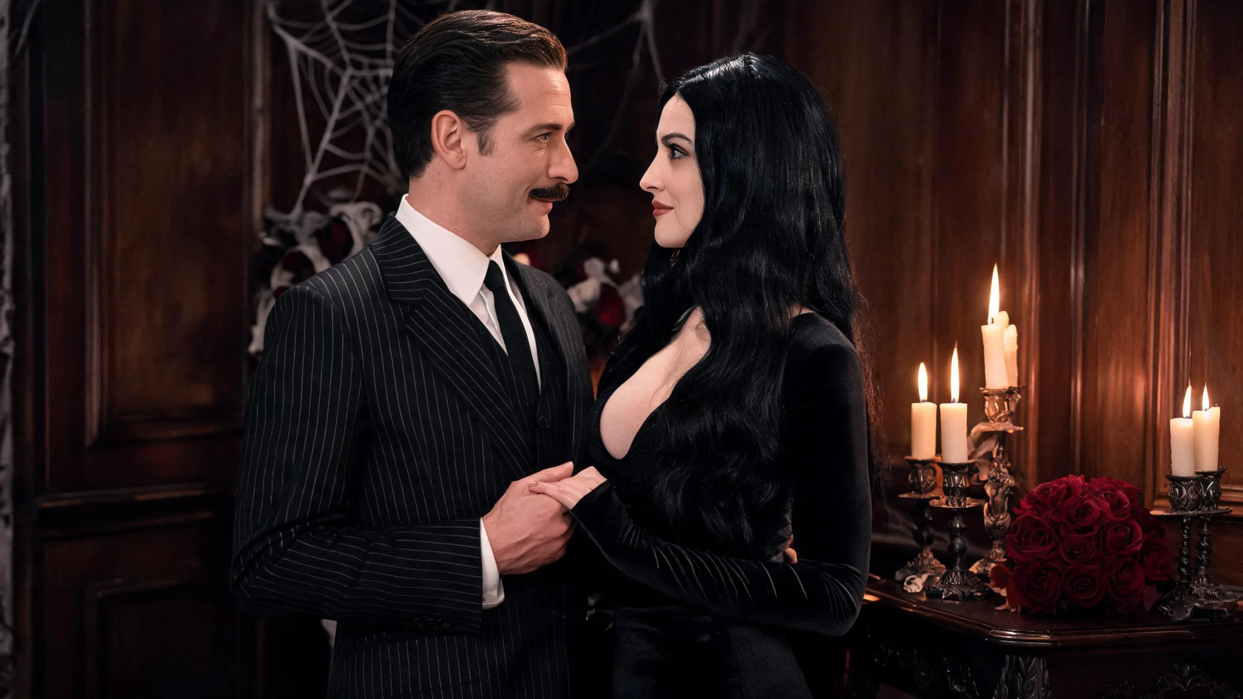 Two male friends in conversation at a Halloween rooftop party at dusk, one styled in a black Gomez Addams-inspired tailored suit, the other in a black leather jacket indie look, warm string lights and other costumed guests interacting in the background