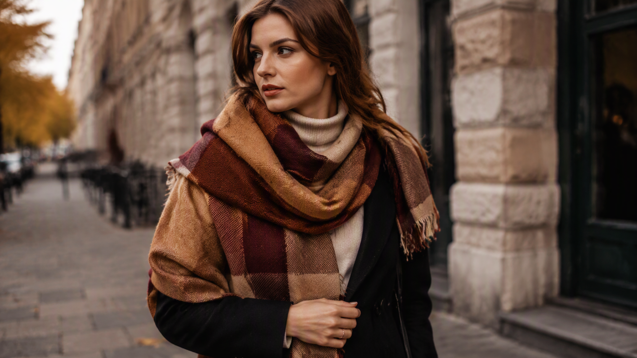 Two cozy fall outfits side by side: a burgundy blanket scarf belted over a black turtleneck and a cream shawl draped over a long wool coat, photographed on a wooden bench with autumn leaves.