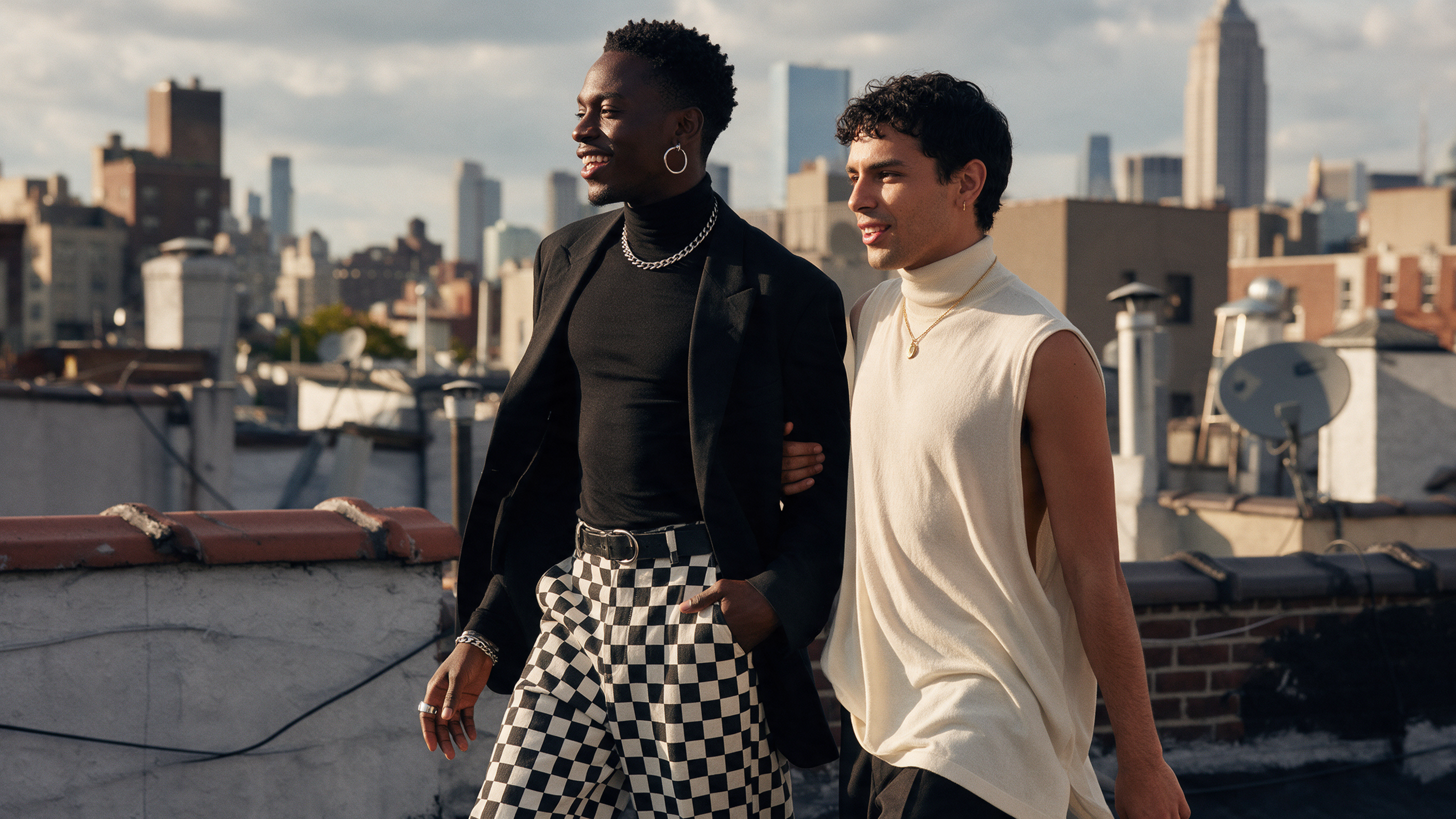 Black man walking through a sunlit city neighborhood wearing an all-black monochrome outfit with wide-leg checkered trousers and silver jewelry, and a Latino man beside him in a flowing sleeveless cream turtleneck with gold necklace.