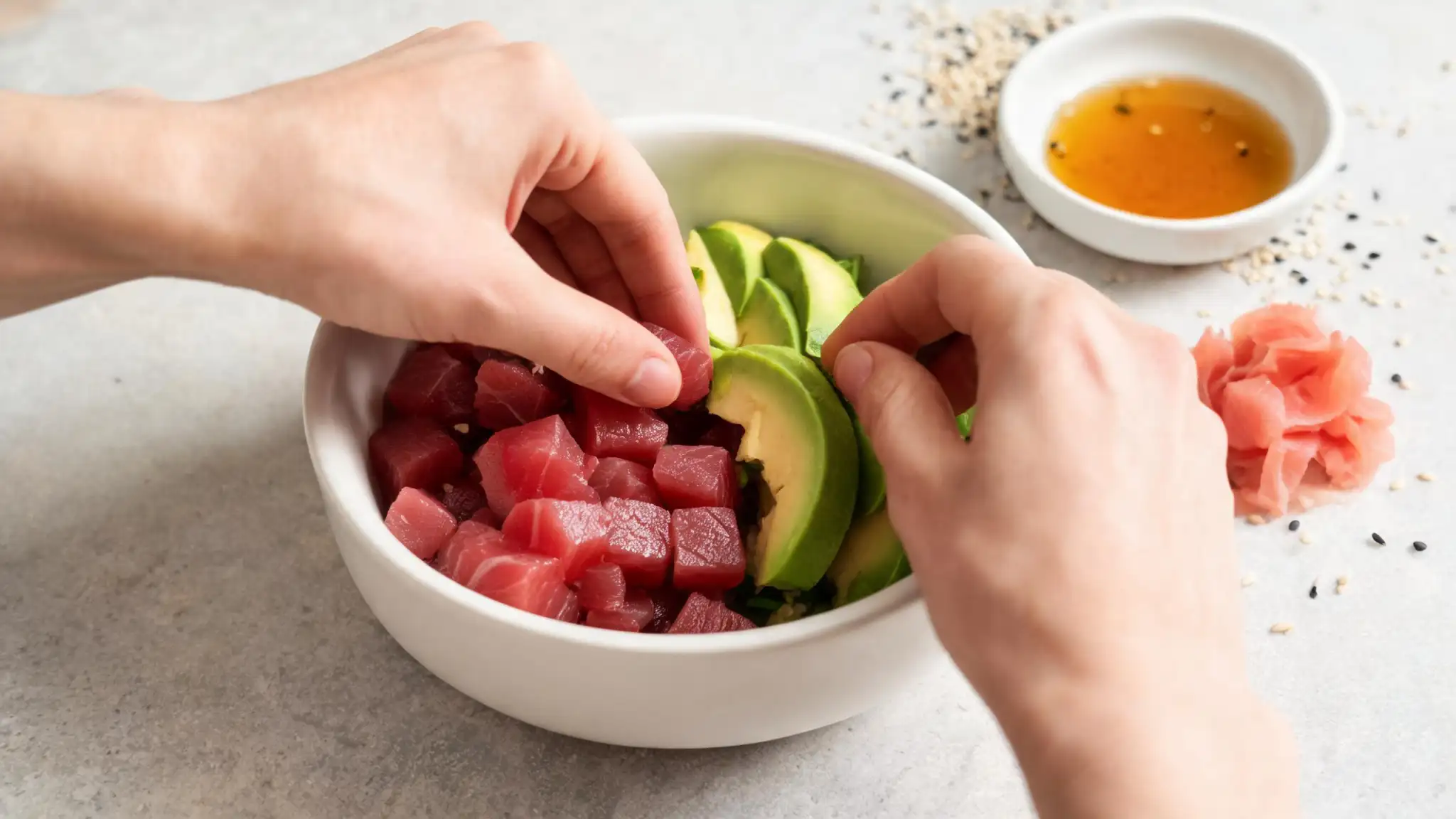 Close-up of hands assembling a fresh poke bowl, adding cubed tuna, sliced avocado, and pickled ginger into a white ceramic bowl, with ponzu sauce and sesame seeds in the background.